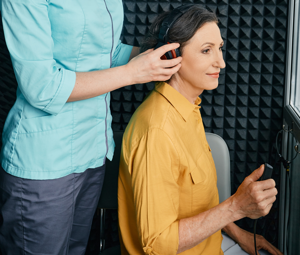 A woman in a yellow shirt sits in a soundproof room holding a button while a healthcare professional places headphones on her for a hearing test.