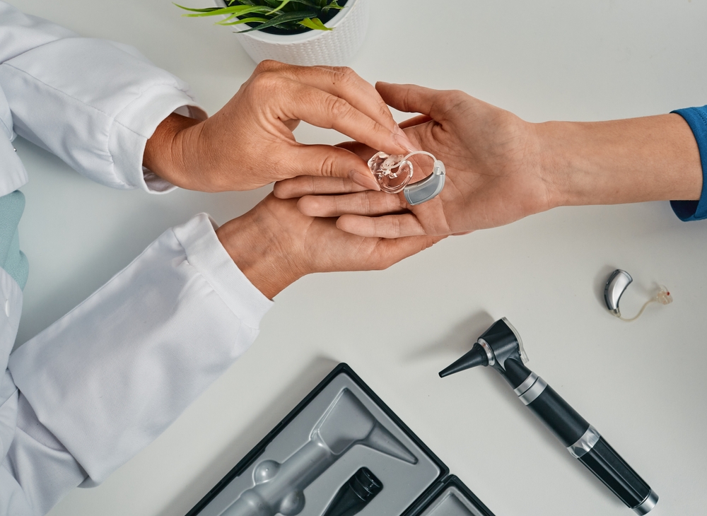 A doctor hands a hearing aid to a patient across a white table, with an otoscope, a hearing aid case, and a potted plant also visible.