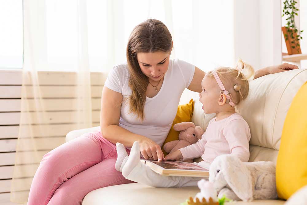 A woman sits on a couch reading a book with a young child who is wearing a cochlear implant and holding a plush toy. The room is bright and cozy with soft furnishings.
