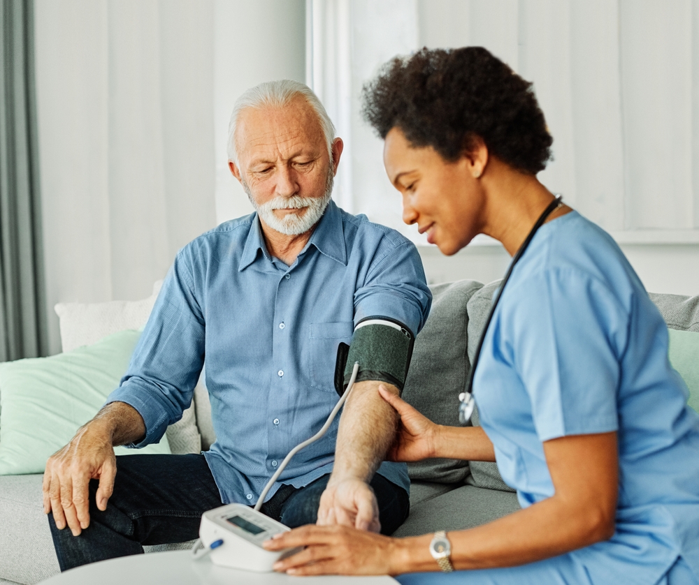 A nurse in blue scrubs checks an older man’s blood pressure using a digital monitor while they sit on a couch in a bright living room.