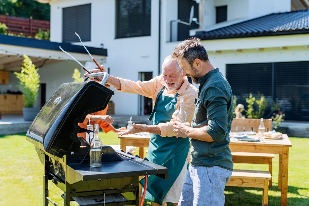 Two men happily grilling food outside in a sunny backyard, standing next to a barbecue grill with a modern house in the background and a wooden table set for a meal nearby.