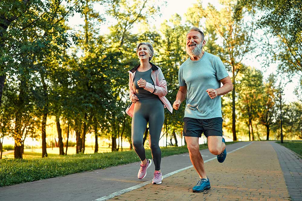 A smiling older couple jogs together on a paved path in a park, surrounded by green trees and bathed in warm sunlight. Both appear energetic and happy, wearing athletic clothing and running shoes.