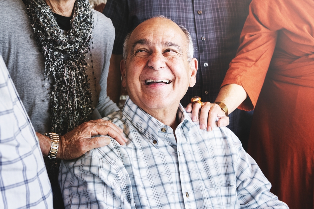 An older man in a plaid shirt smiles warmly while surrounded by friends, who gently rest their hands on his shoulders and arms in a supportive, joyful group setting.