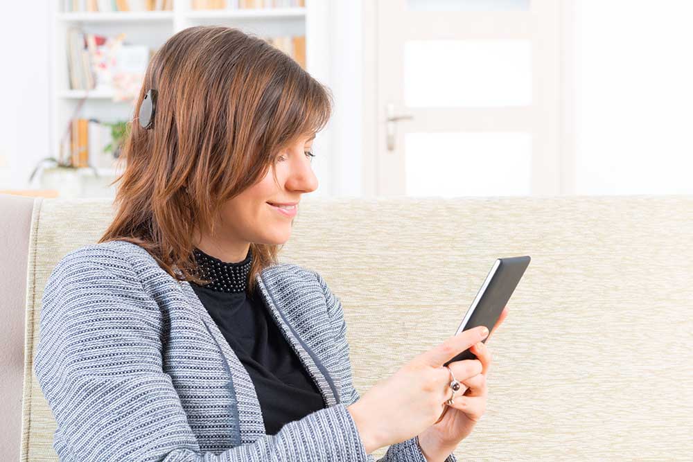 A woman with a cochlear implant sits on a couch, smiling while using a smartphone or tablet. She is wearing a gray sweater and appears relaxed in a bright, cozy room.