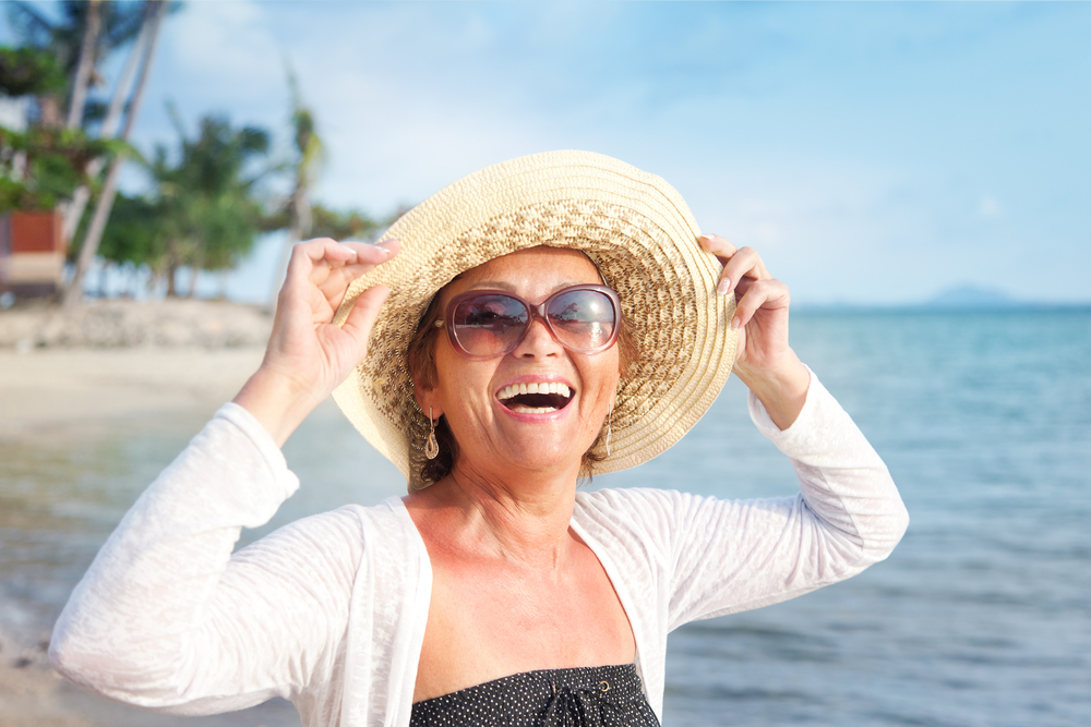 Smiling woman wearing sunglasses and a sunhat stands on a tropical beach near the water, enjoying a sunny day. Palm trees and blue sky are visible in the background.