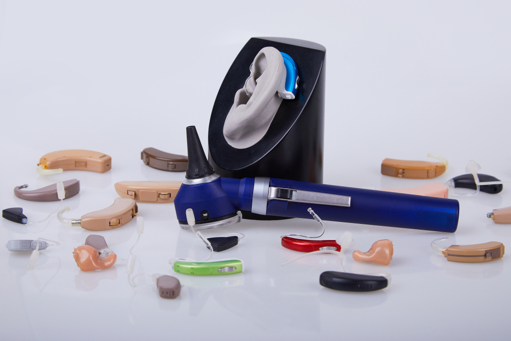 A blue otoscope is placed among various hearing aids of different colors and styles, with a model ear and hearing aid on a black stand in the background, all set against a white surface.
