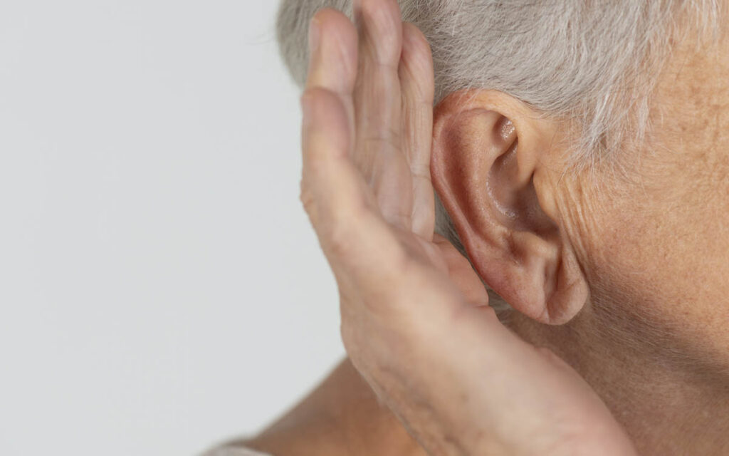 Close-up of an older adult’s ear, with their hand cupped behind it, as if trying to hear better. The background is plain and light-colored.