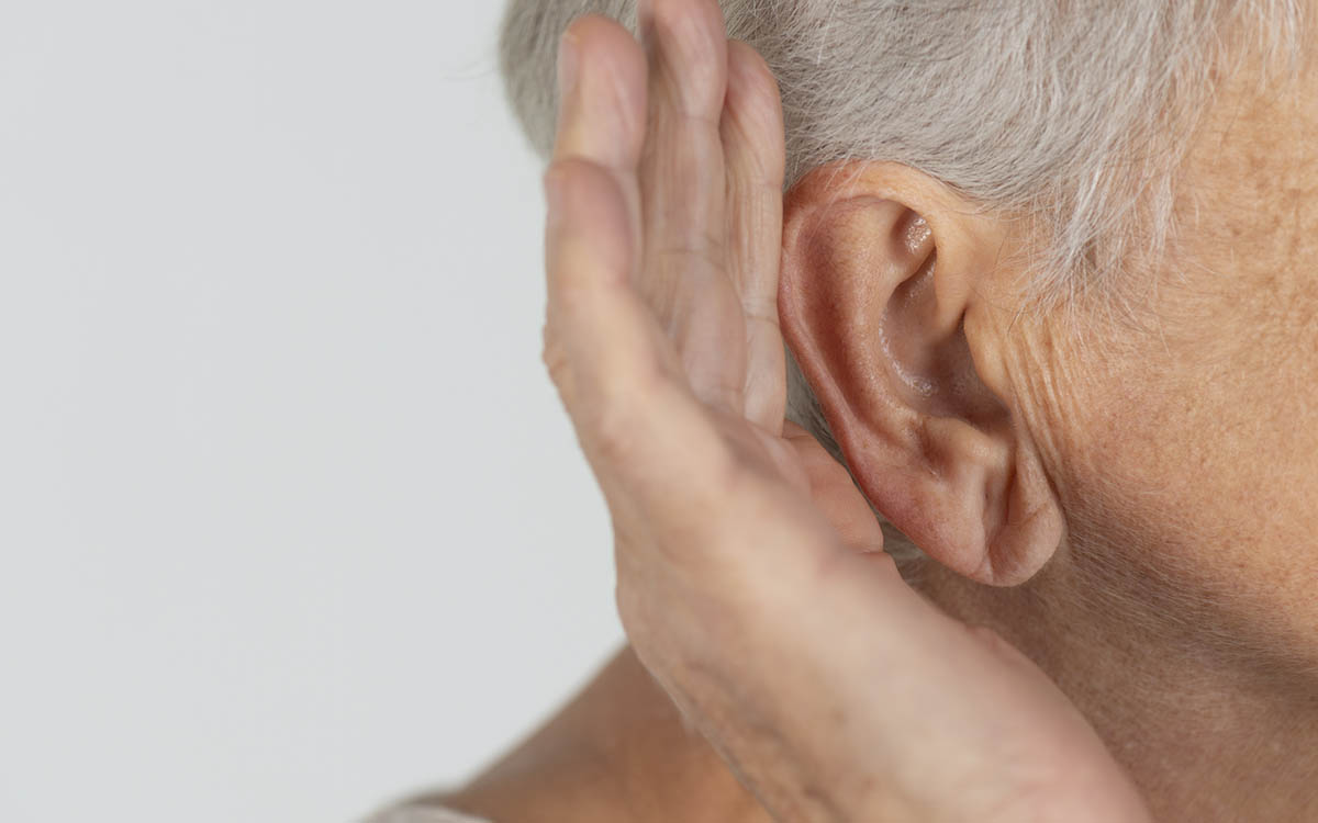 Close-up of an older adult’s ear, with their hand cupped behind it, as if trying to hear better. The background is plain and light-colored.
