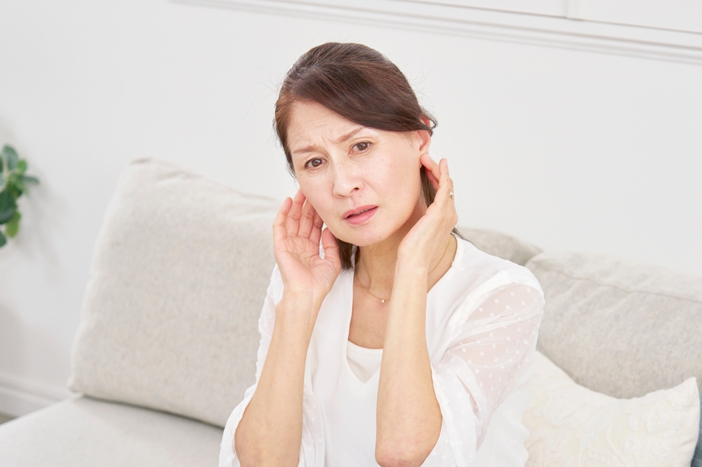 A middle-aged woman with a concerned expression sits on a sofa, touching both of her ears as if experiencing discomfort or pain. She is wearing a white blouse in a bright, softly lit room.