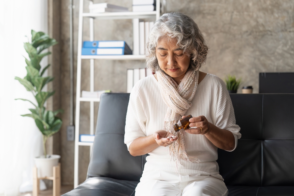 An older woman with gray hair sits on a couch, looking down as she pours pills from a brown bottle into her hand. She wears a light scarf and white clothing, with a bookshelf and plant in the background.