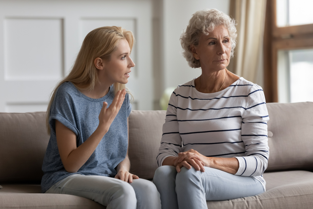A young woman talks emotionally to an older woman sitting beside her on a couch. The older woman looks away with a serious expression, while the younger woman gestures with her hand, appearing frustrated.