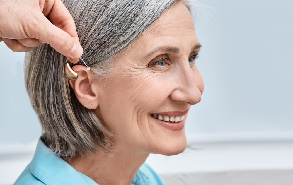 Smiling older woman with gray hair is having a hearing aid placed behind her ear by another persons hand. She is wearing a light blue shirt and appears content.