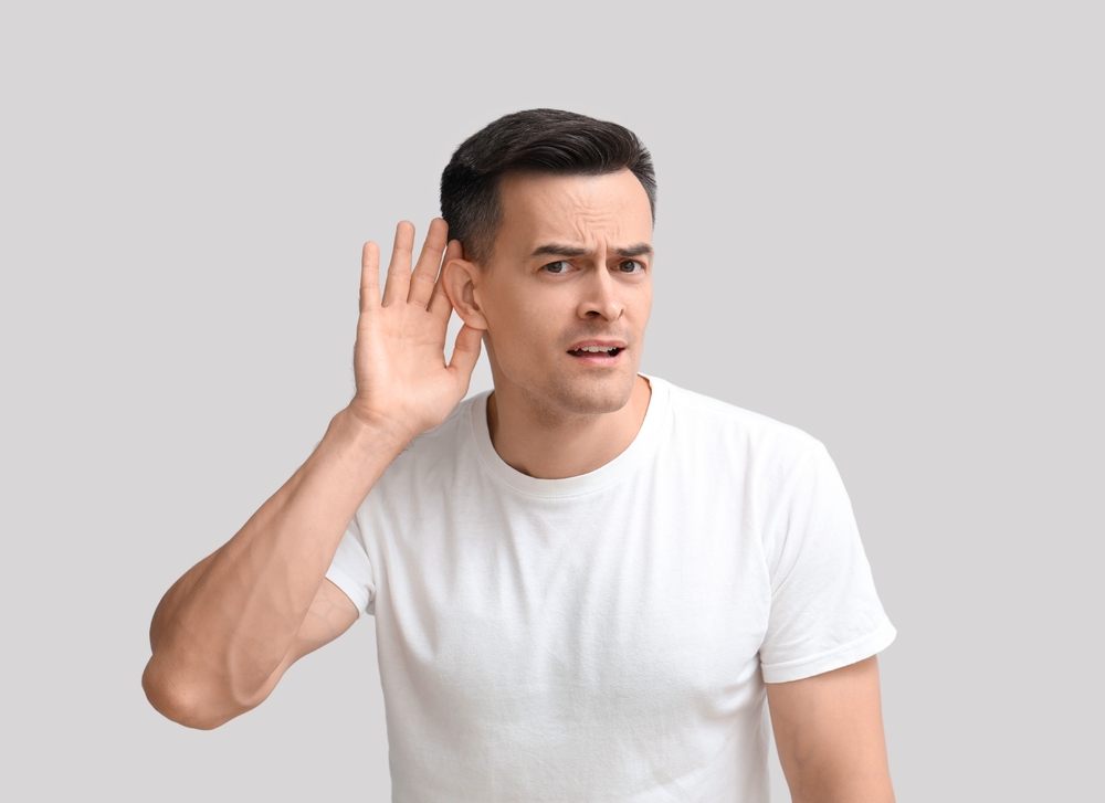 A man in a white t-shirt holds his hand to his ear and looks confused, as if trying to hear something better, against a plain gray background.