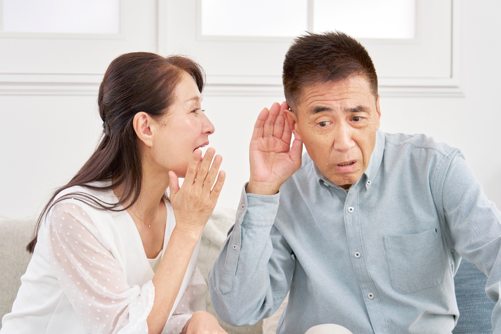 A woman whispers into the ear of a man who is leaning in and cupping his ear, appearing to have difficulty hearing her. They are sitting together indoors on a sofa.