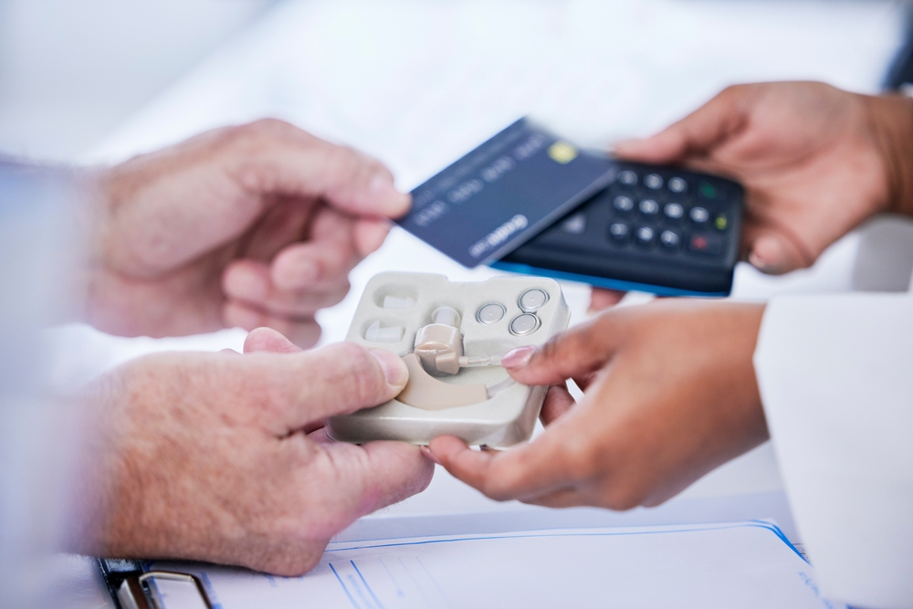 A person pays with a credit card using a card reader while receiving a hearing aid device in a case from a healthcare professional.