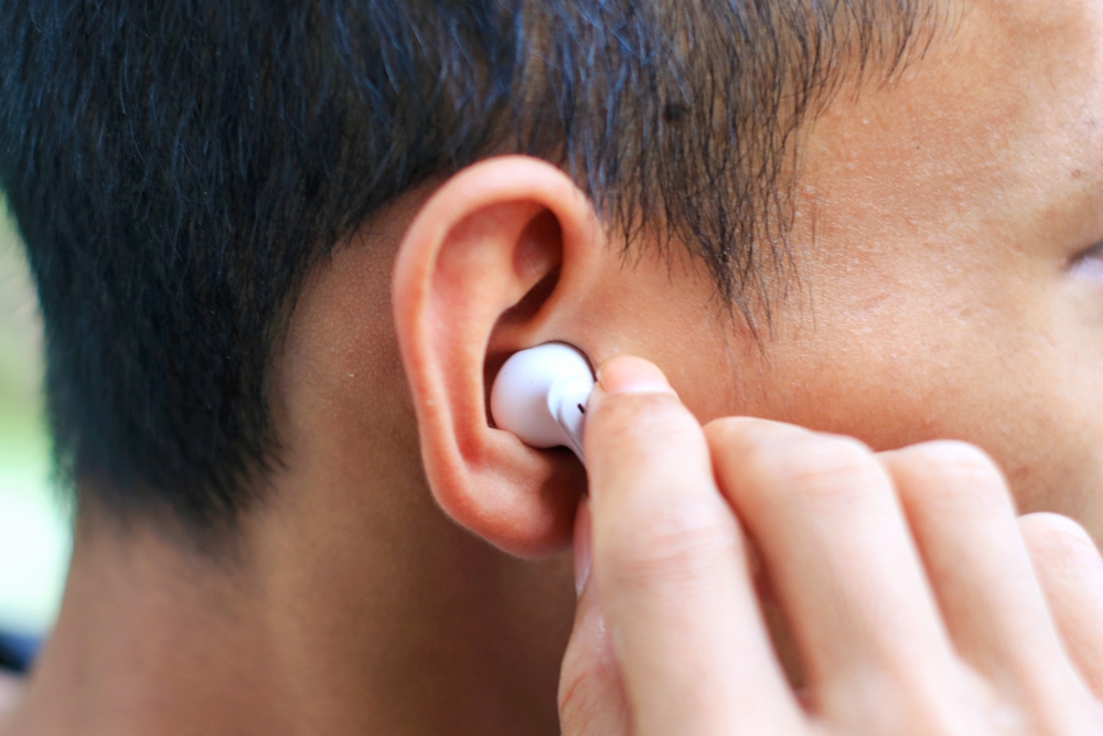 A person is inserting a white wireless earbud into their right ear, with their hand holding the earbud close to the ear. The focus is on the ear and the earbud.