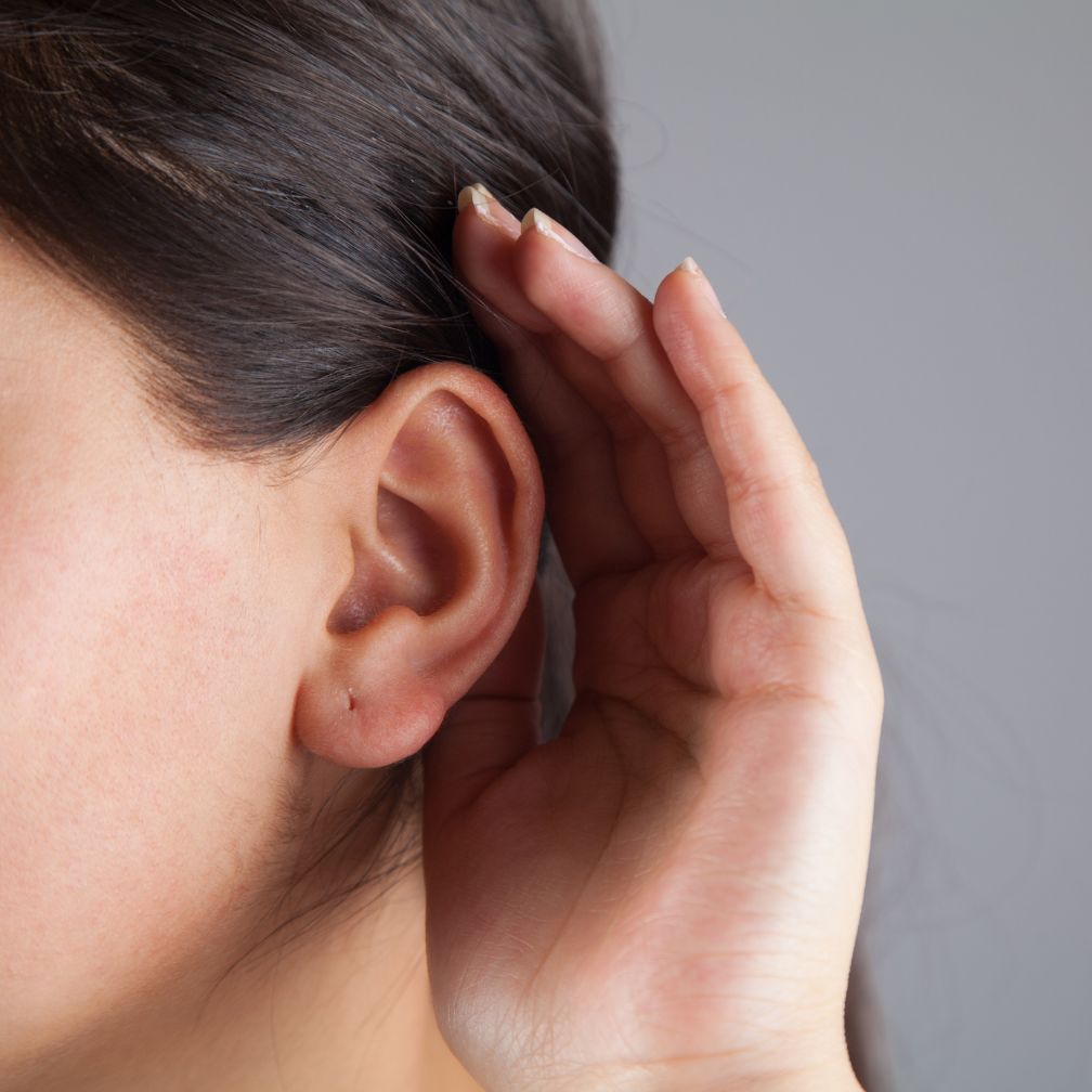 A person holding their hand up to their ear, as if trying to hear better. The image shows the side of their face, ear, and hand against a neutral background.