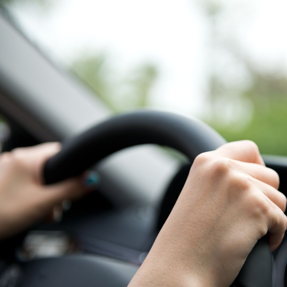 Close-up of a persons hands gripping a car steering wheel, with a blurred view of the car interior and greenery visible through the window in the background.