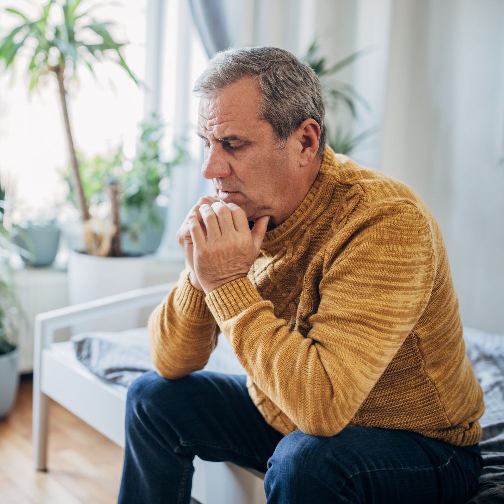 An older man in a mustard sweater sits on a bed, looking thoughtful and concerned, with his hands clasped near his chin. There are several potted plants in the background.