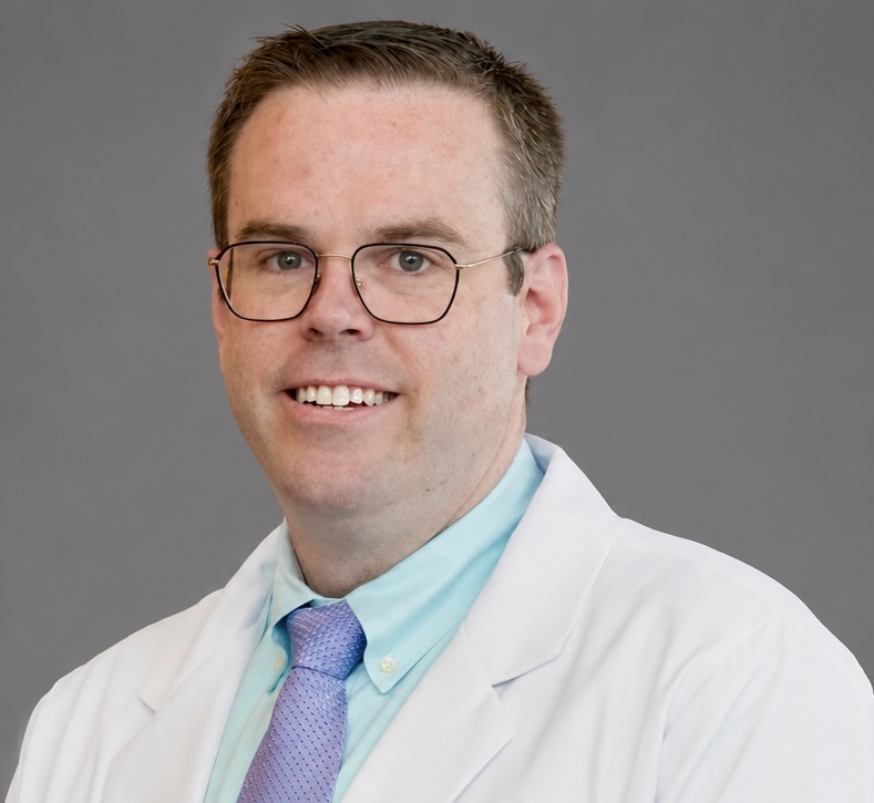 A man wearing glasses, a white lab coat, a light blue shirt, and a lavender tie smiles in front of a plain gray background.