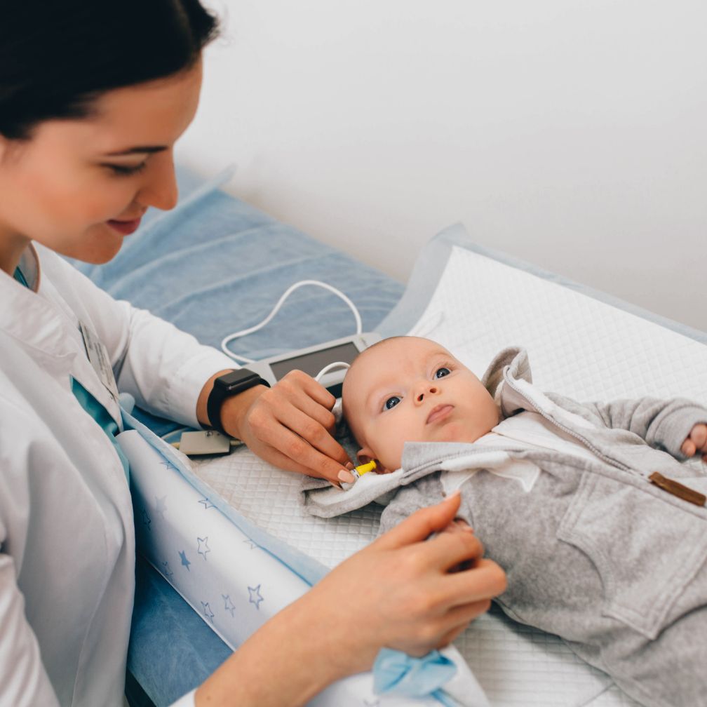 A healthcare professional tests a babys hearing using a small device while the baby lies on an exam table, looking up and wearing a gray hoodie.