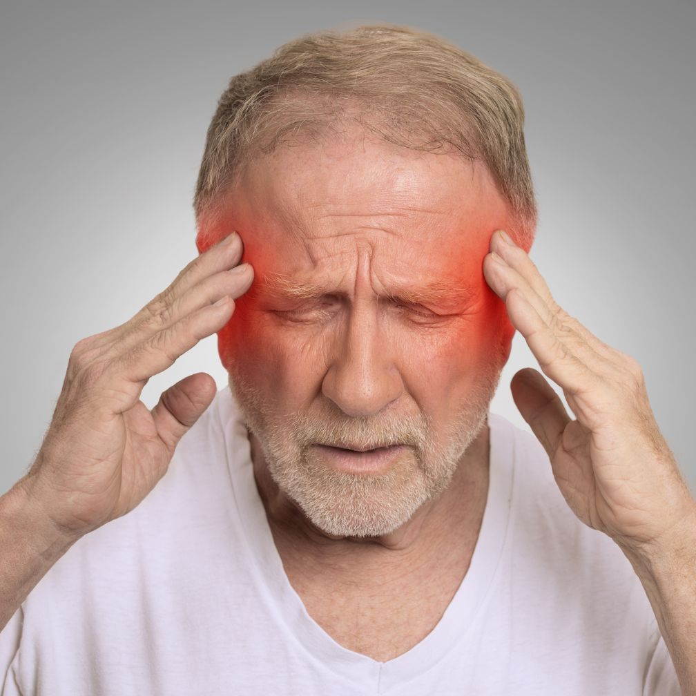 An older man with closed eyes touches his temples, showing pain. Red highlights on his temples suggest a headache or migraine.