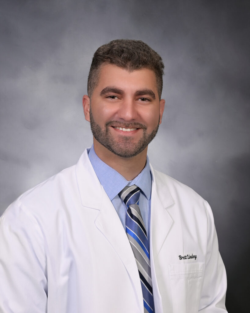 A man with short brown hair and a beard smiles while wearing a white lab coat, blue shirt, and striped tie, posing in front of a gray background.
