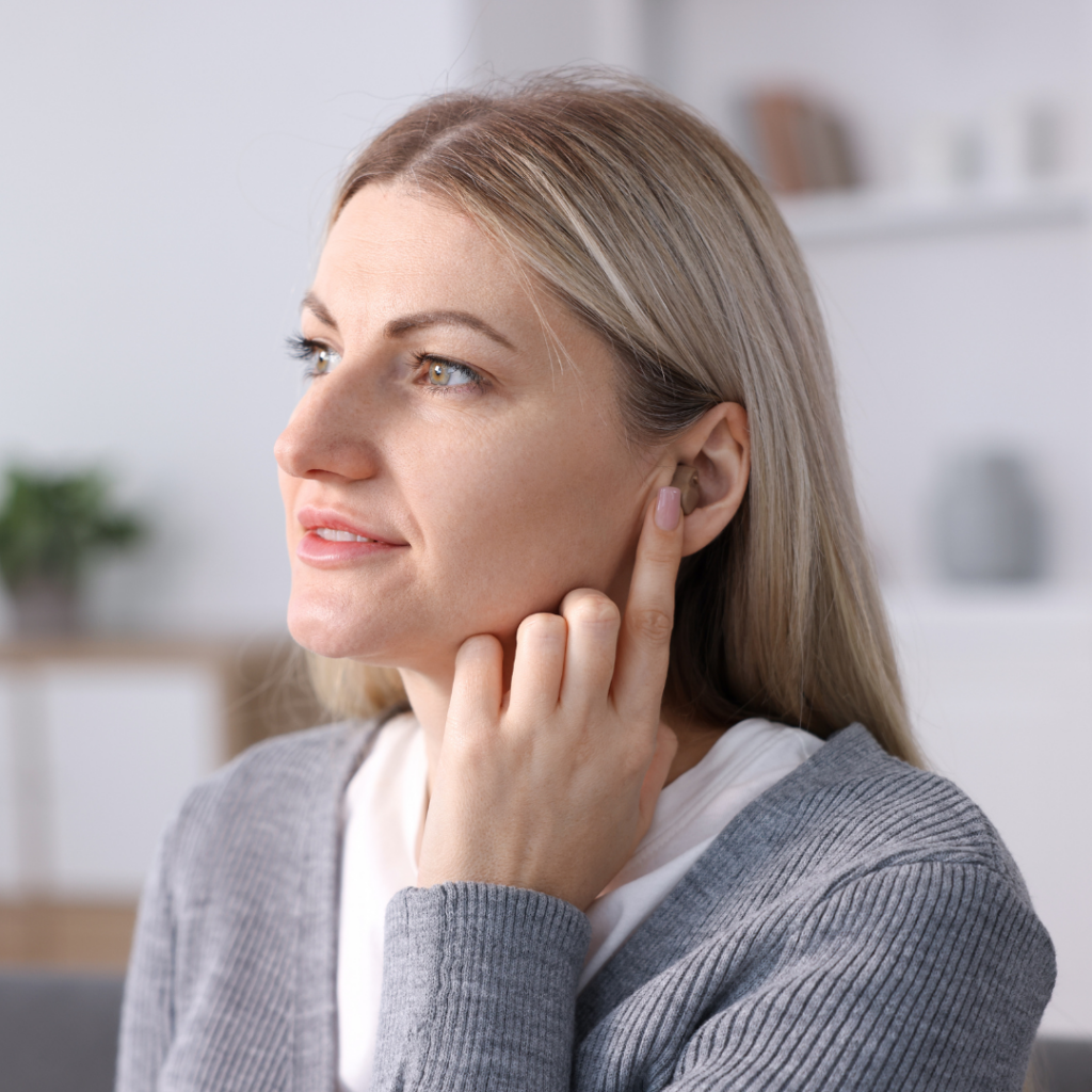 A woman with long blonde hair touches her ear with her finger, appearing thoughtful. She wears a gray sweater and sits indoors with blurred shelves and plants in the background.