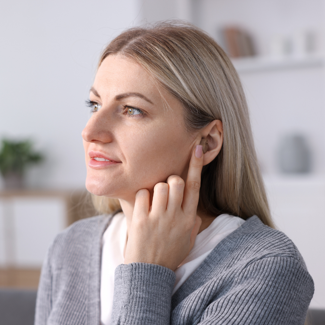 A woman with long blonde hair touches her ear with her finger, appearing thoughtful. She wears a gray sweater and sits indoors with blurred shelves and plants in the background.